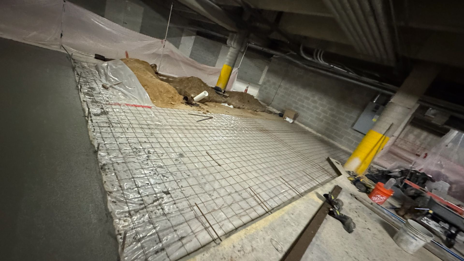 A construction site interior showing a metal wire mesh laid over soil and plastic sheeting, prepared for a concrete pour.