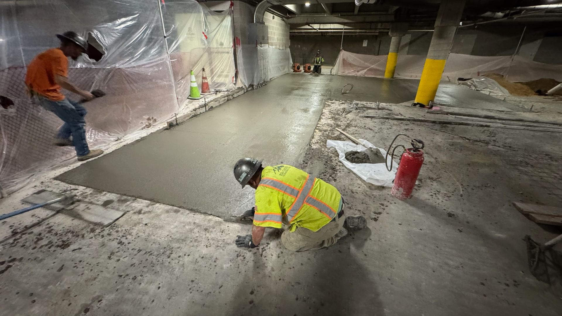 Two workers in high-visibility safety gear smooth wet concrete in an indoor construction site with concrete pillars.