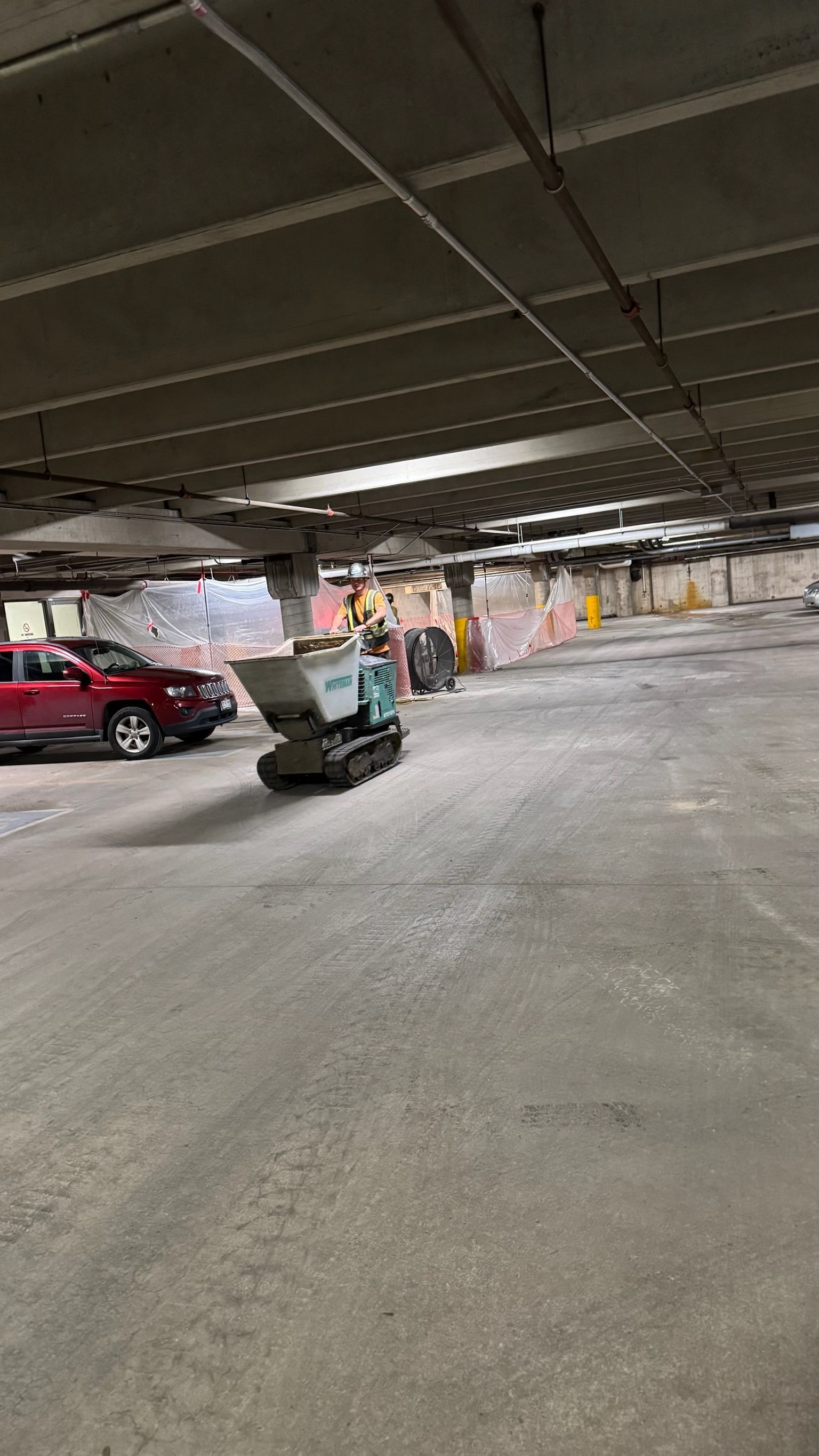 A small, teal tracked dumper moving through a dimly lit indoor parking garage near a red vehicle and construction fencing.