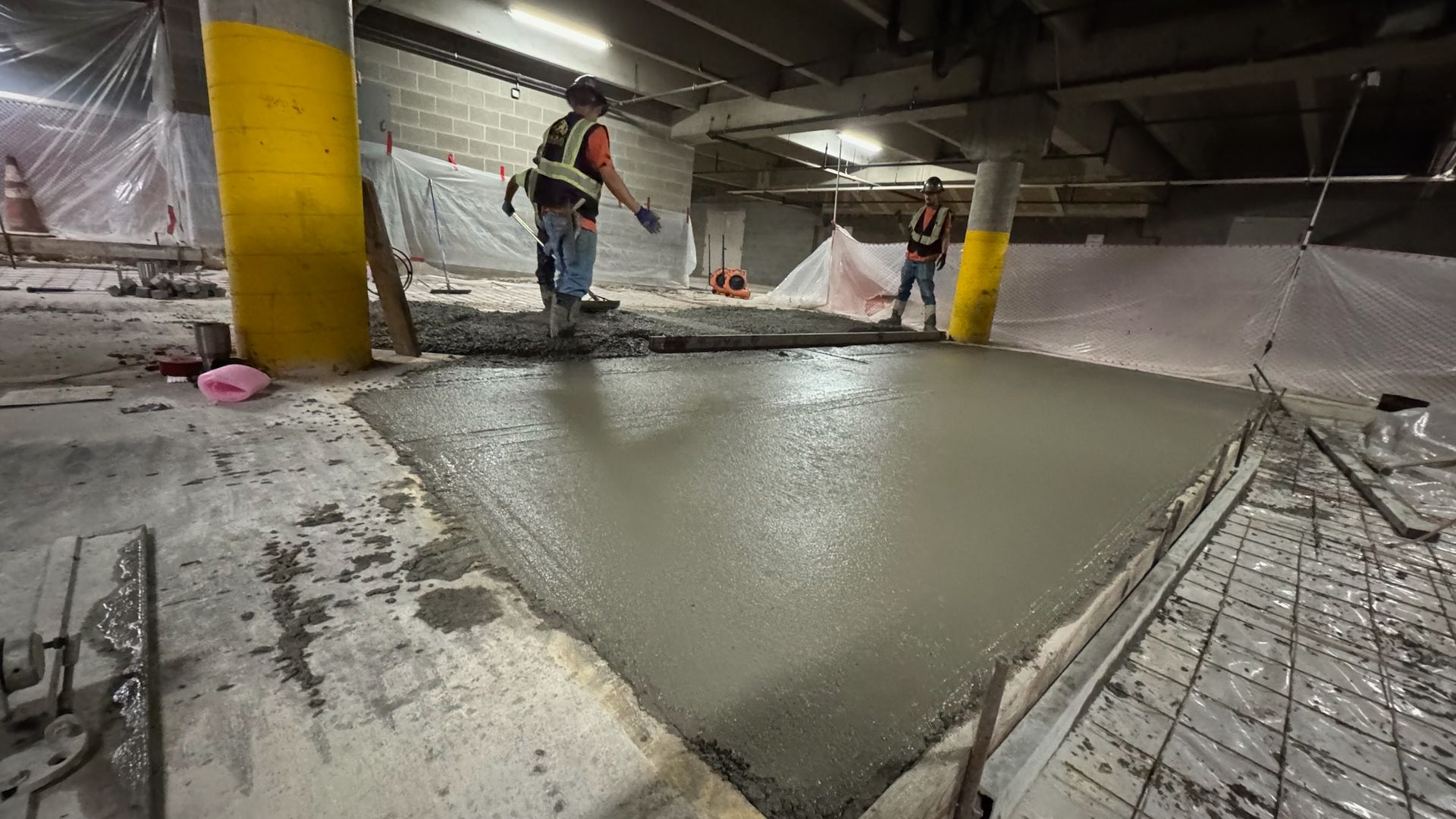 Two workers in high-visibility vests spread wet concrete onto a floor in an indoor construction site with yellow columns.