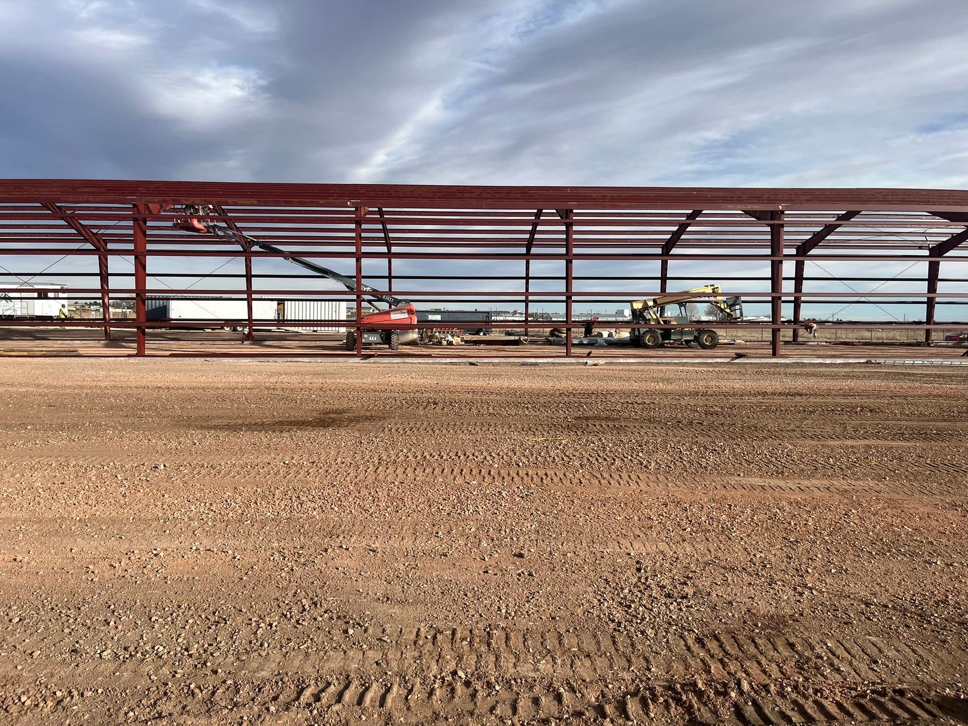 Construction of a long, red-steel frame building on a wide, dirt-covered site under a cloudy sky.