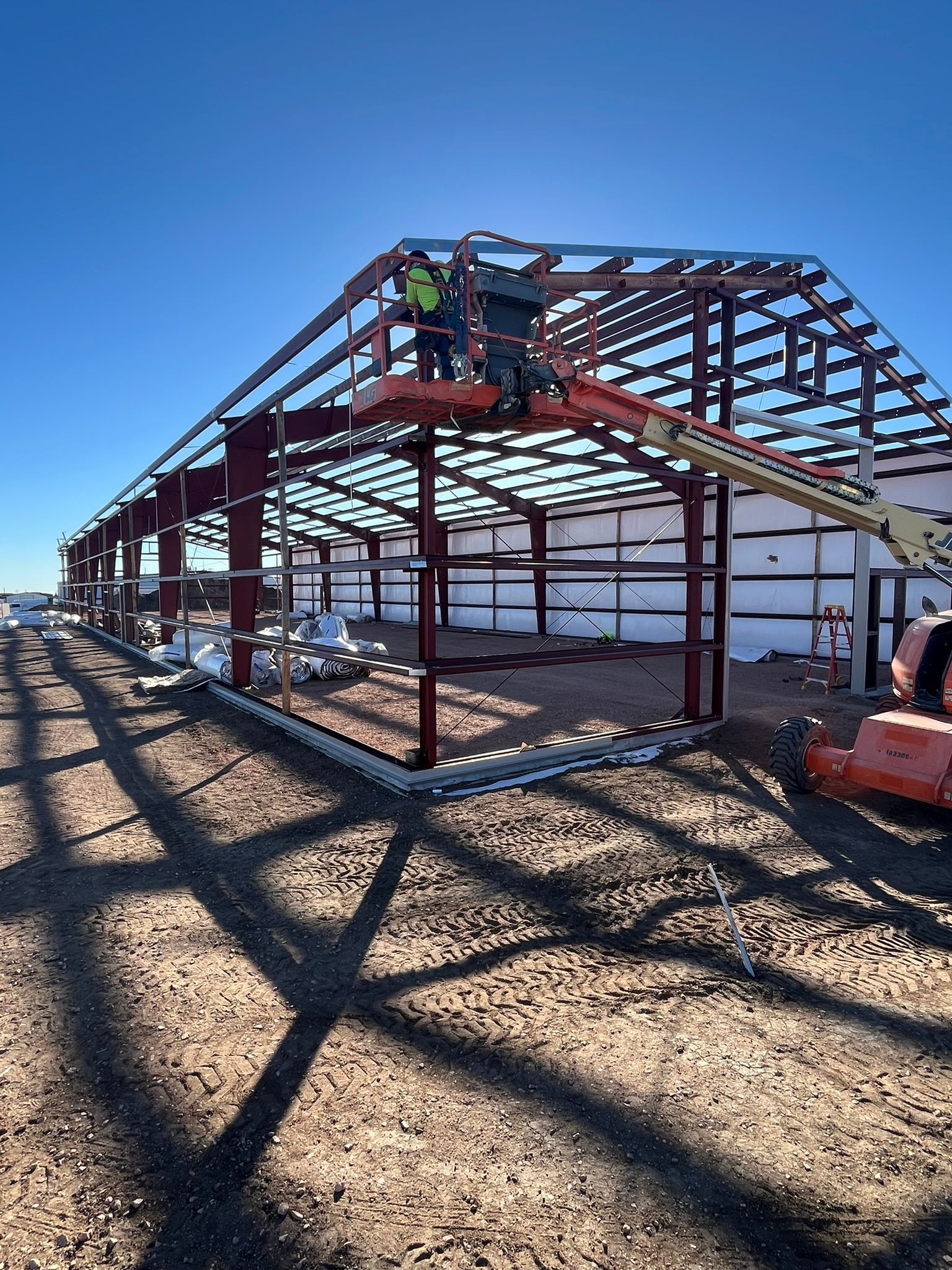 A worker on a boom lift installs steel roof purlins on the red metal frame of an unfinished building under a blue sky.