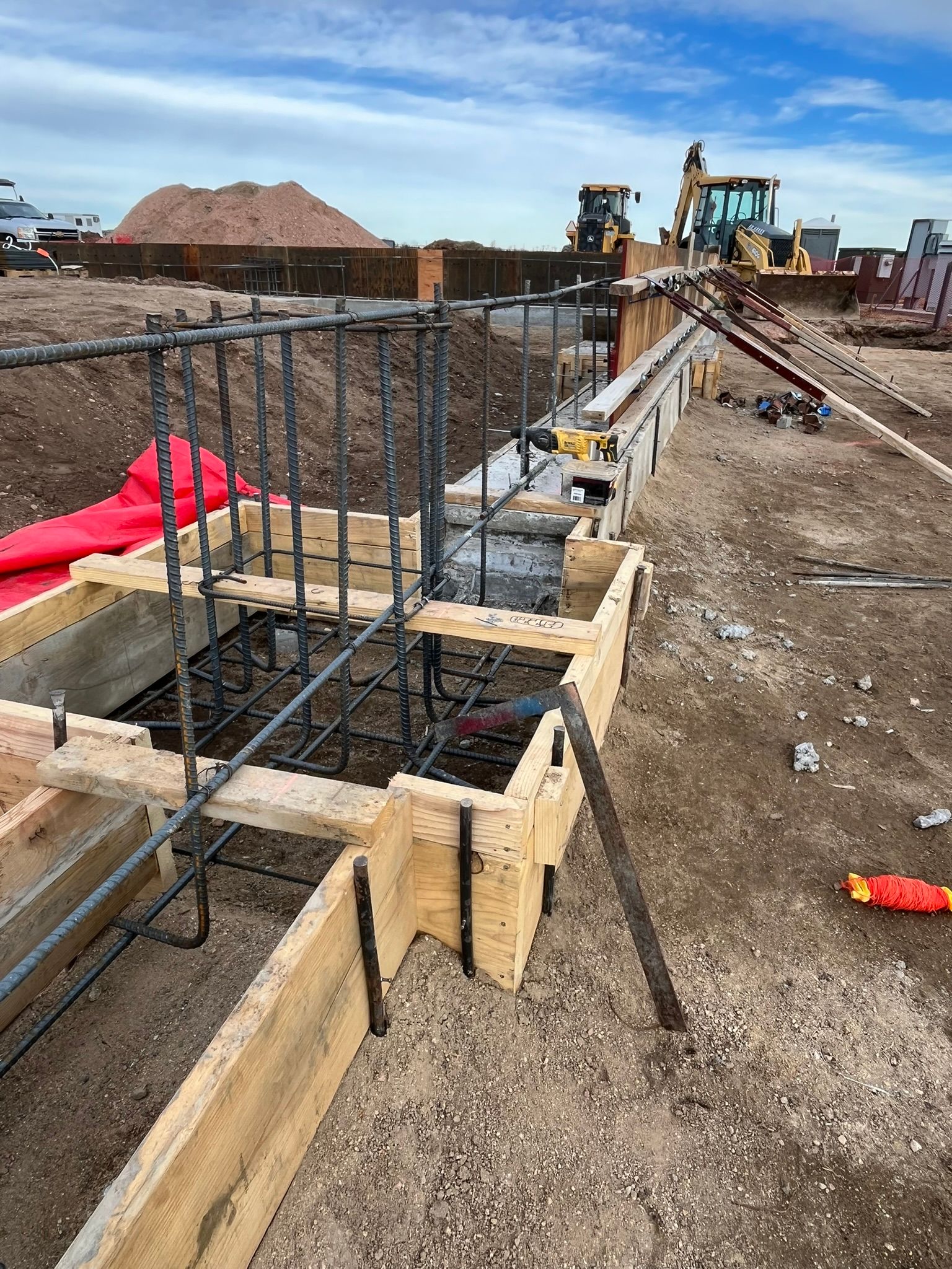 Construction site with wooden concrete forms, metal rebar cages, and heavy machinery in the background.