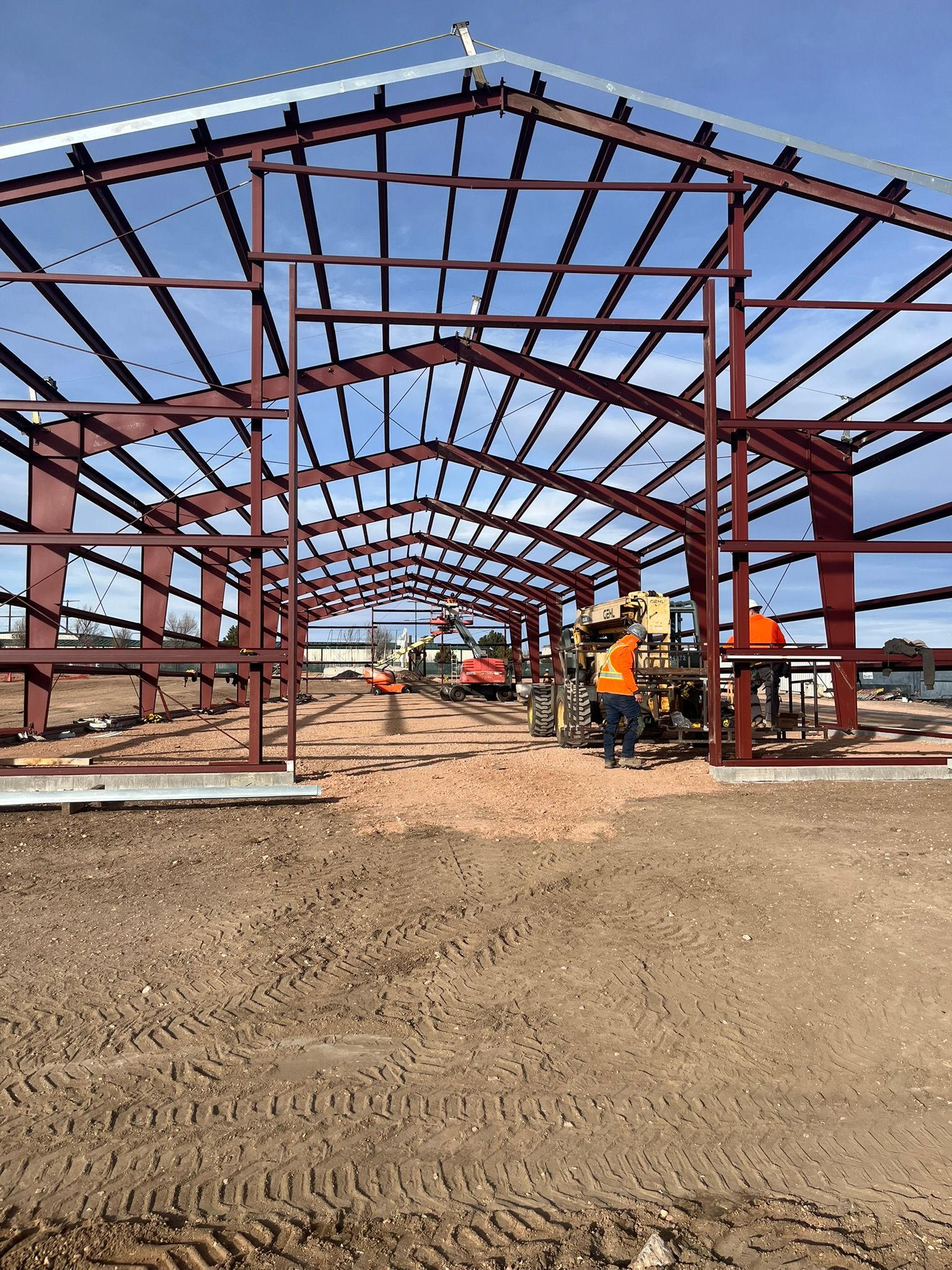 Construction workers stand under the red steel frame of a large building structure on a dirt lot under a blue sky.