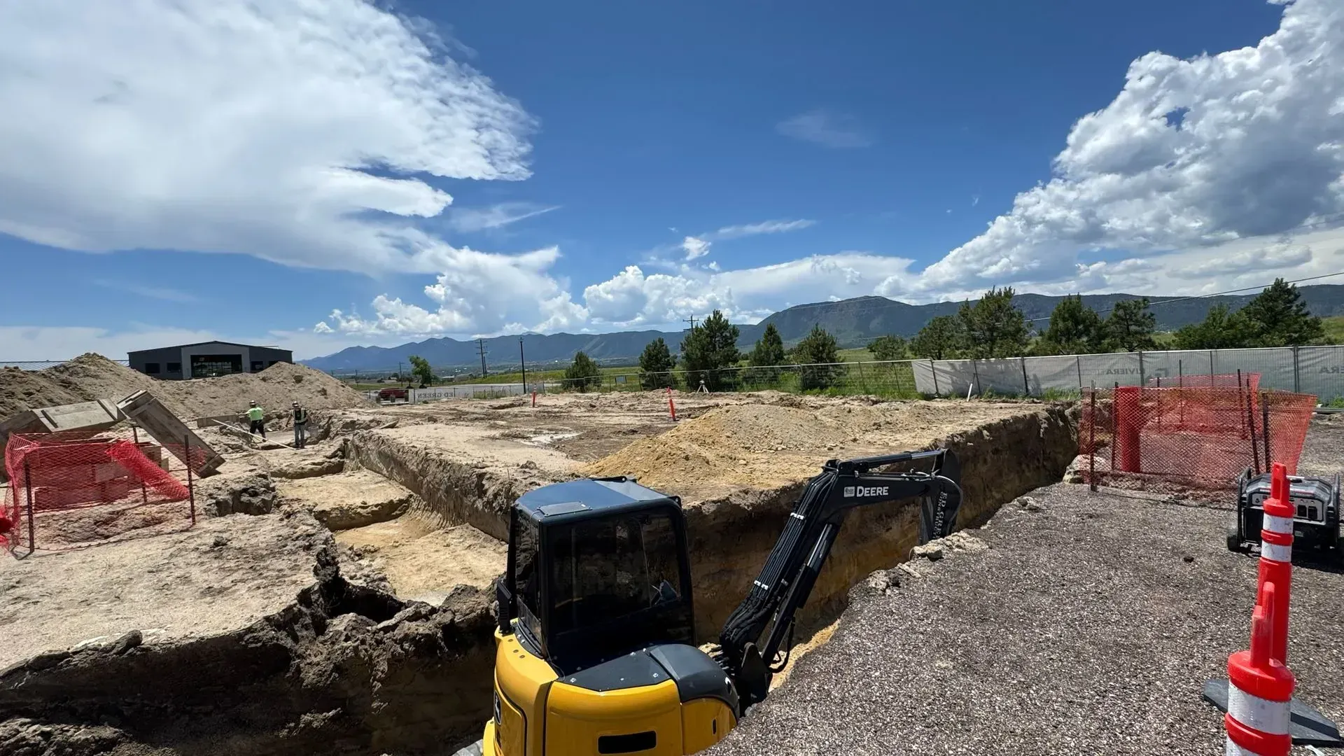 A yellow excavator sits in a dirt foundation trench at a construction site with mountains under a cloudy blue sky.