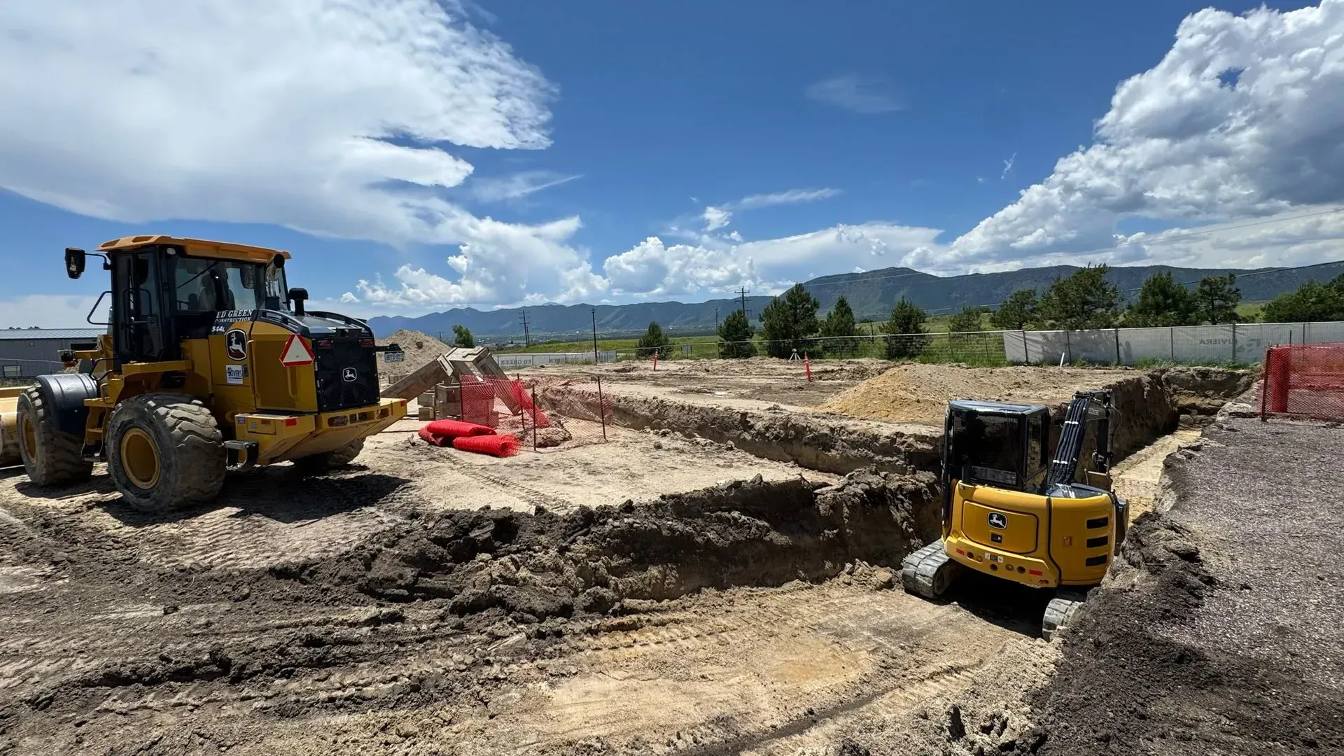 A yellow front loader and a compact excavator work at a construction site under a blue, cloudy sky.