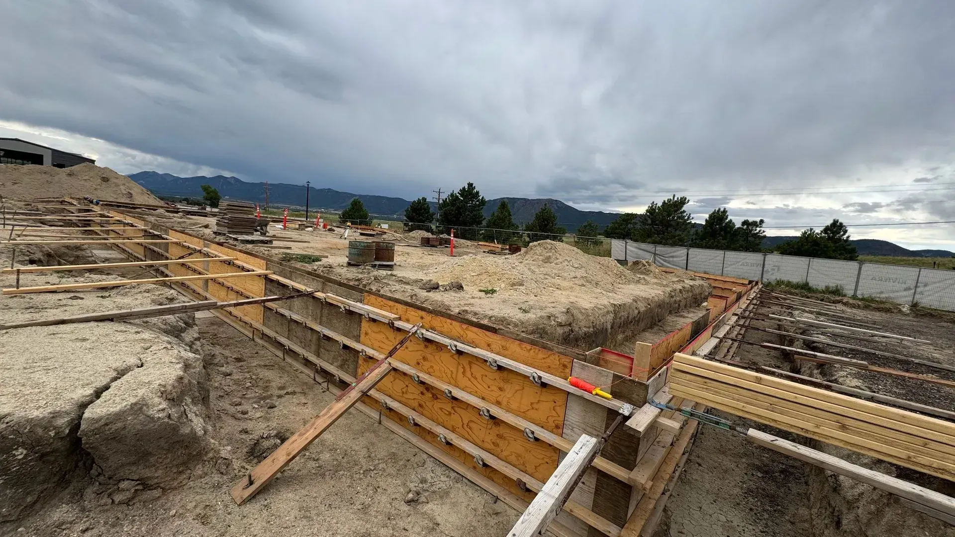 Construction site showing wooden foundation forms set in an excavated dirt lot under a cloudy sky.