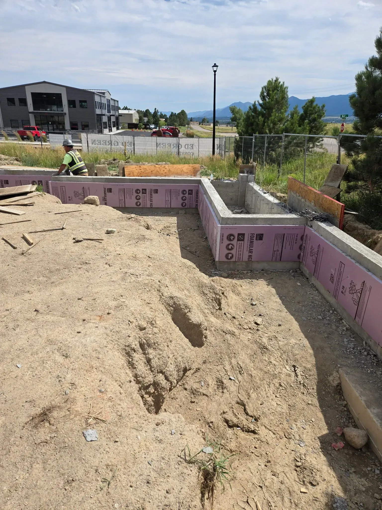 A construction site with a worker standing near concrete foundation walls insulated with pink foam panels.