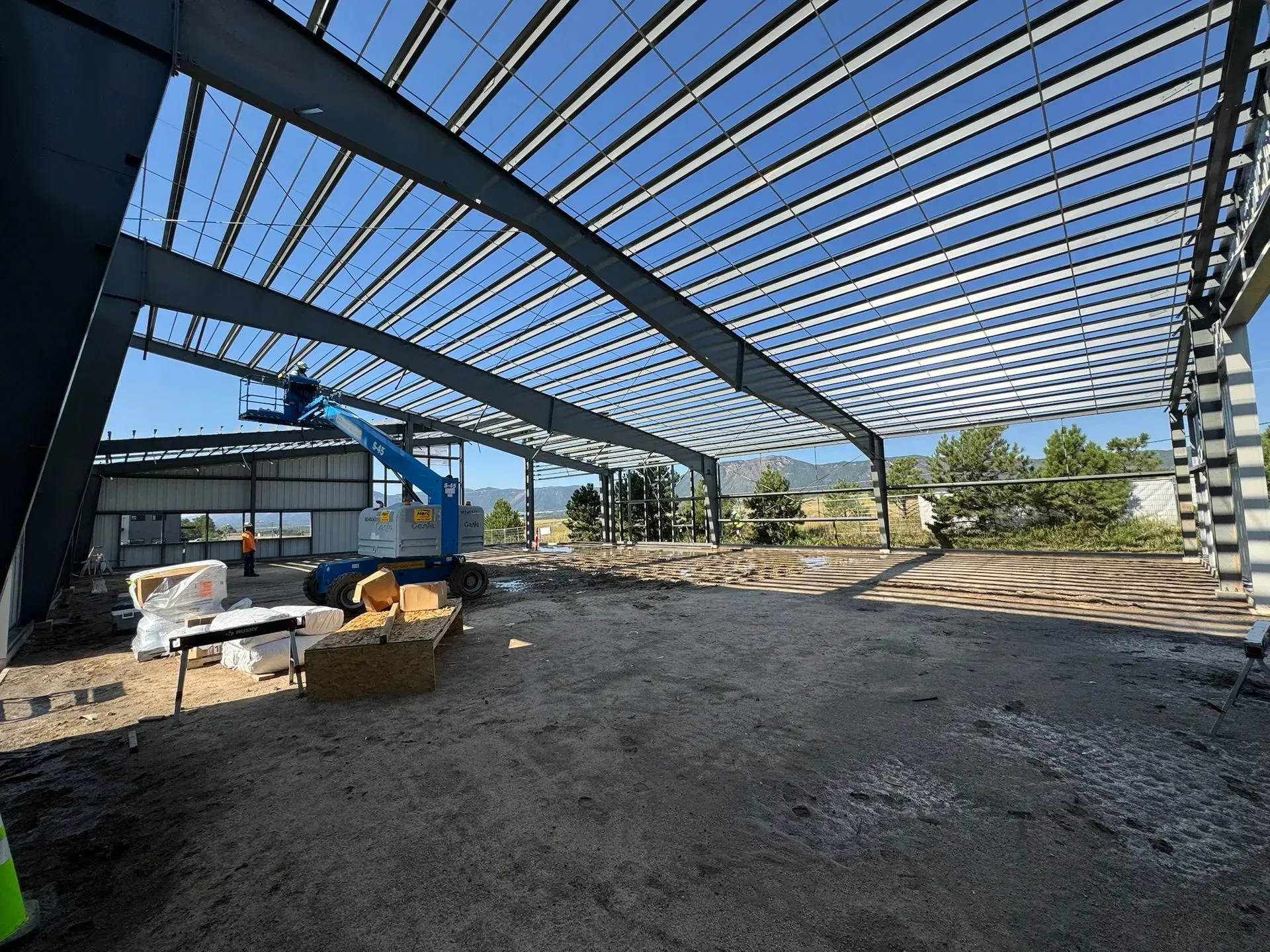 Construction site showing a large steel beam frame and roof structure under a blue sky, with a blue aerial lift parked.