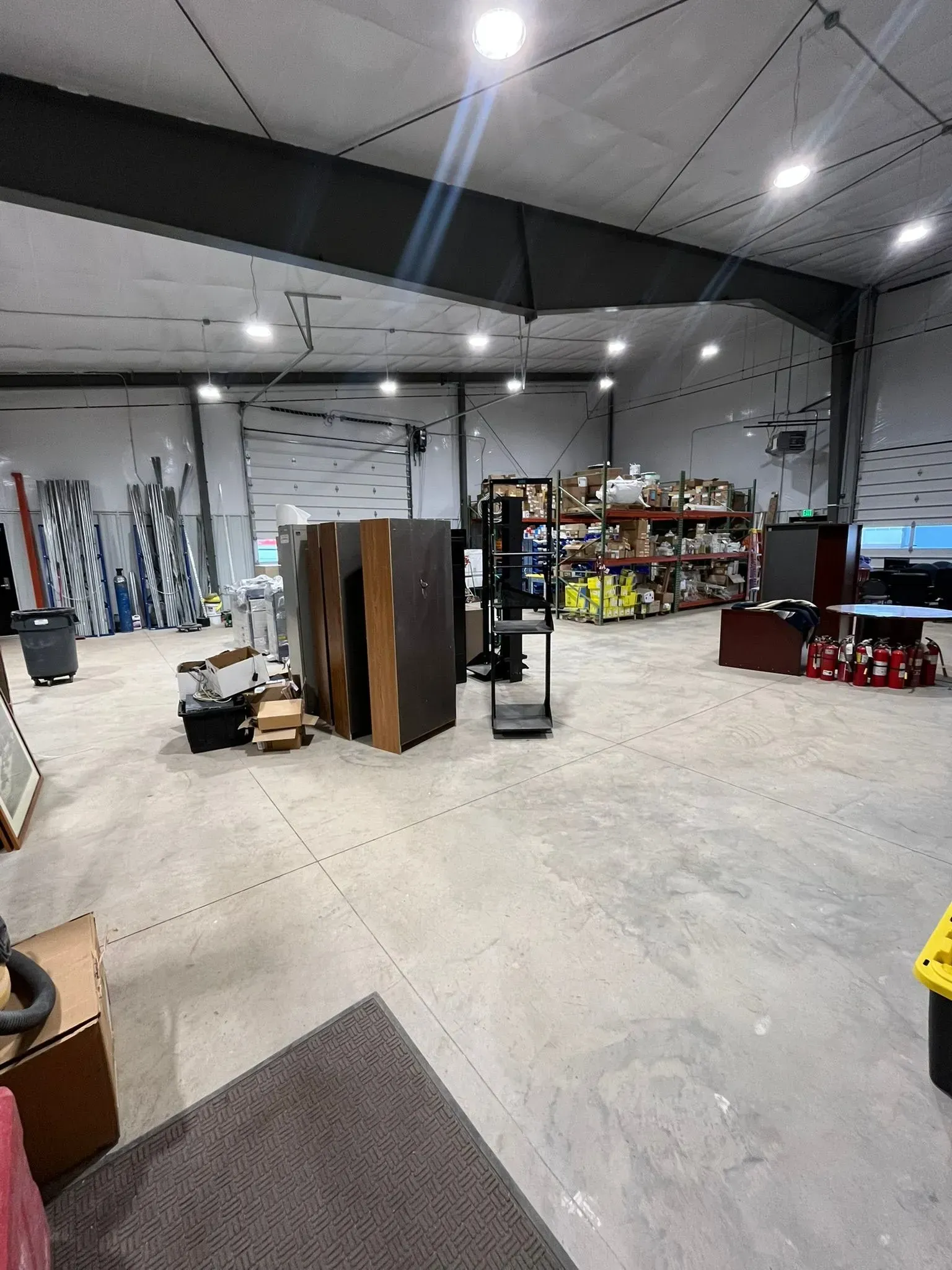 A wide, indoor view of a warehouse with concrete floors, featuring shelving units, stacked materials, and overhead lighting.