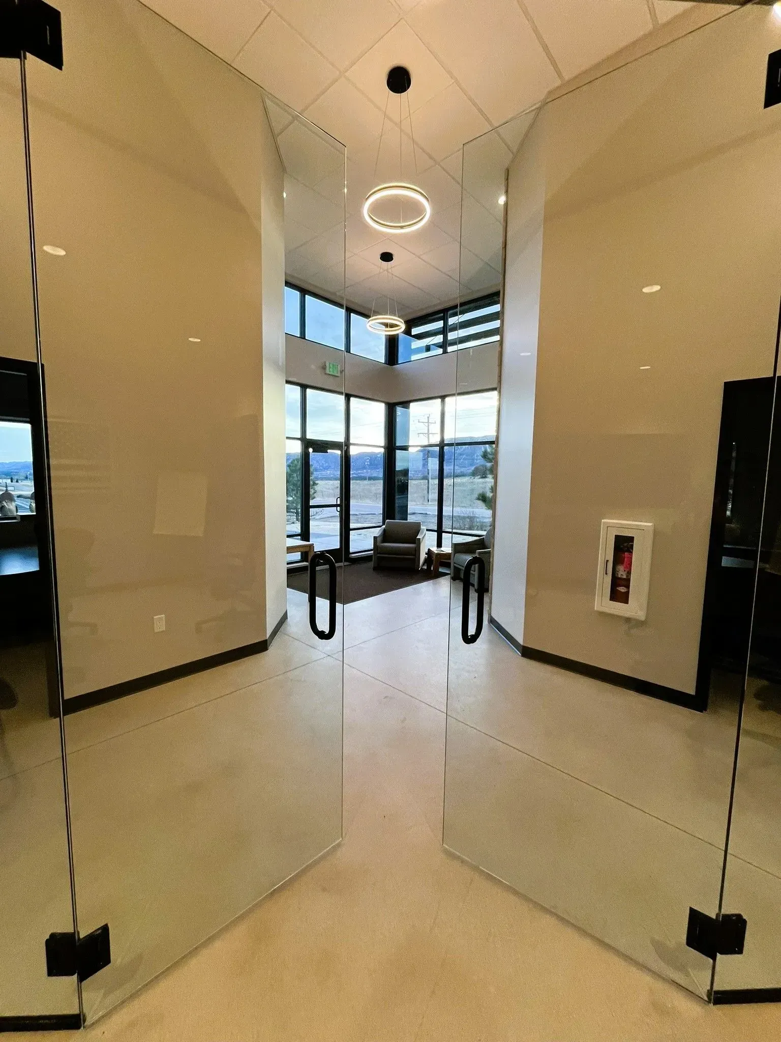 A brightly lit office entryway with open glass doors leading to a lobby area with large windows, seating, and a fire alarm.
