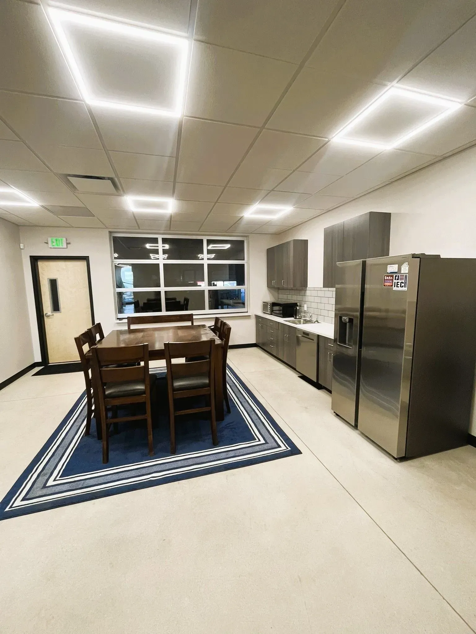 A breakroom featuring a dining table on a patterned rug, a stainless steel refrigerator, and cabinets against the wall.