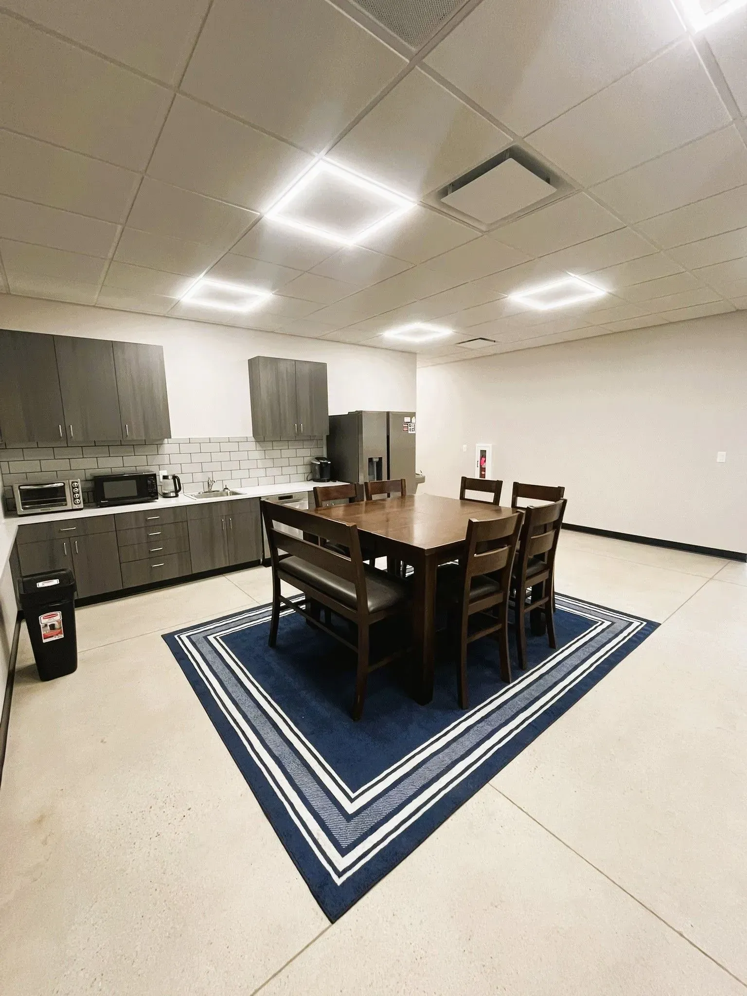 An office breakroom with a square wooden table, chairs, and bench on a blue-and-white patterned rug, plus gray cabinets.