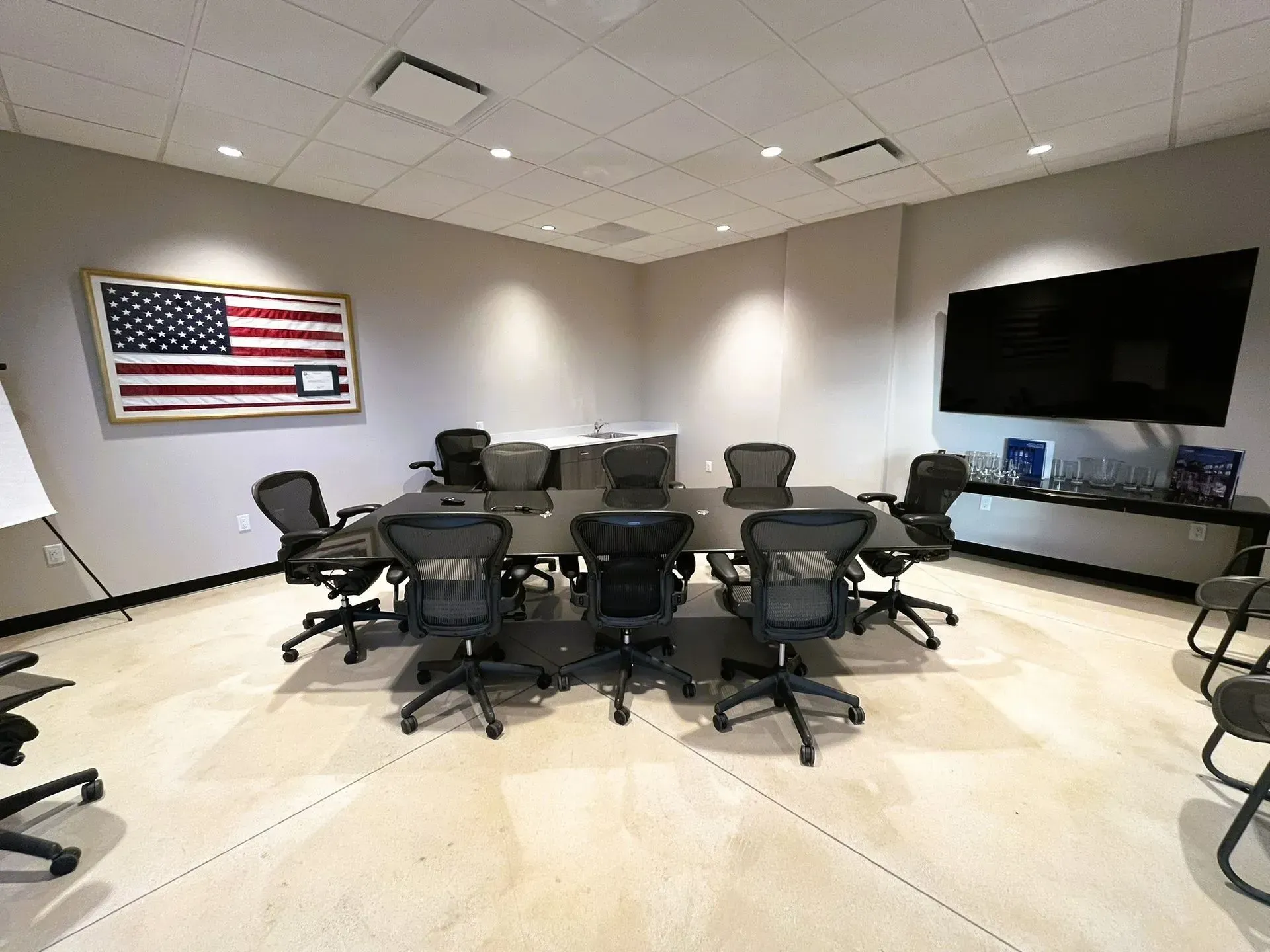 A modern conference room with a long table, ergonomic chairs, a wall-mounted US flag artwork, and a large flat-screen TV.