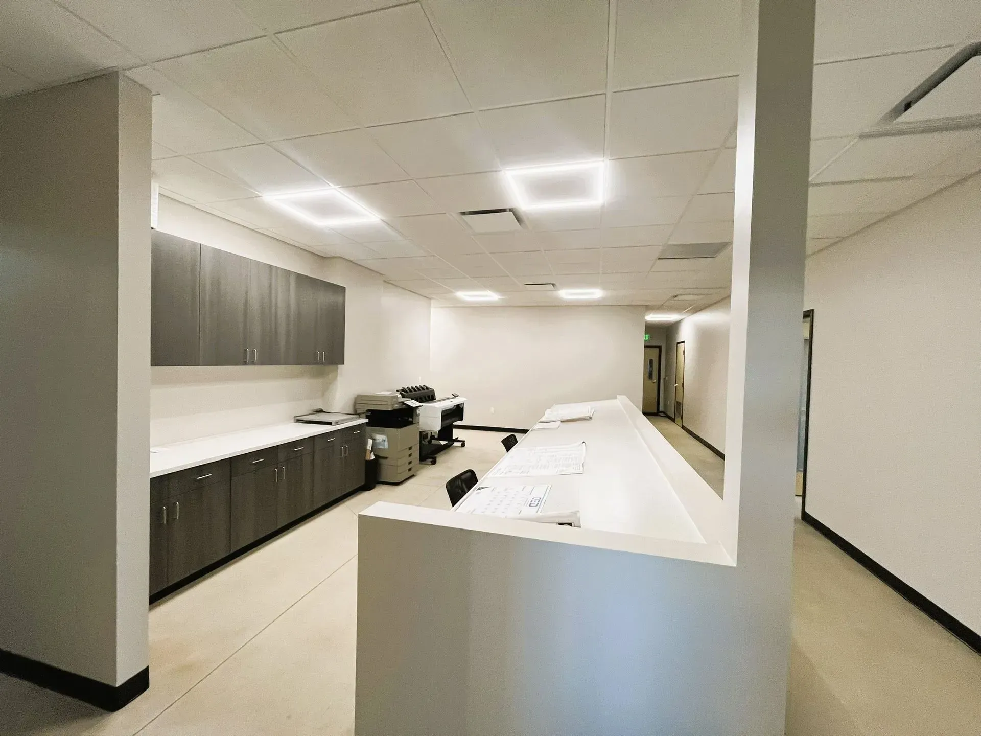 A bright, modern office space featuring dark grey cabinetry, a long white counter, and a printer on a light tiled floor.