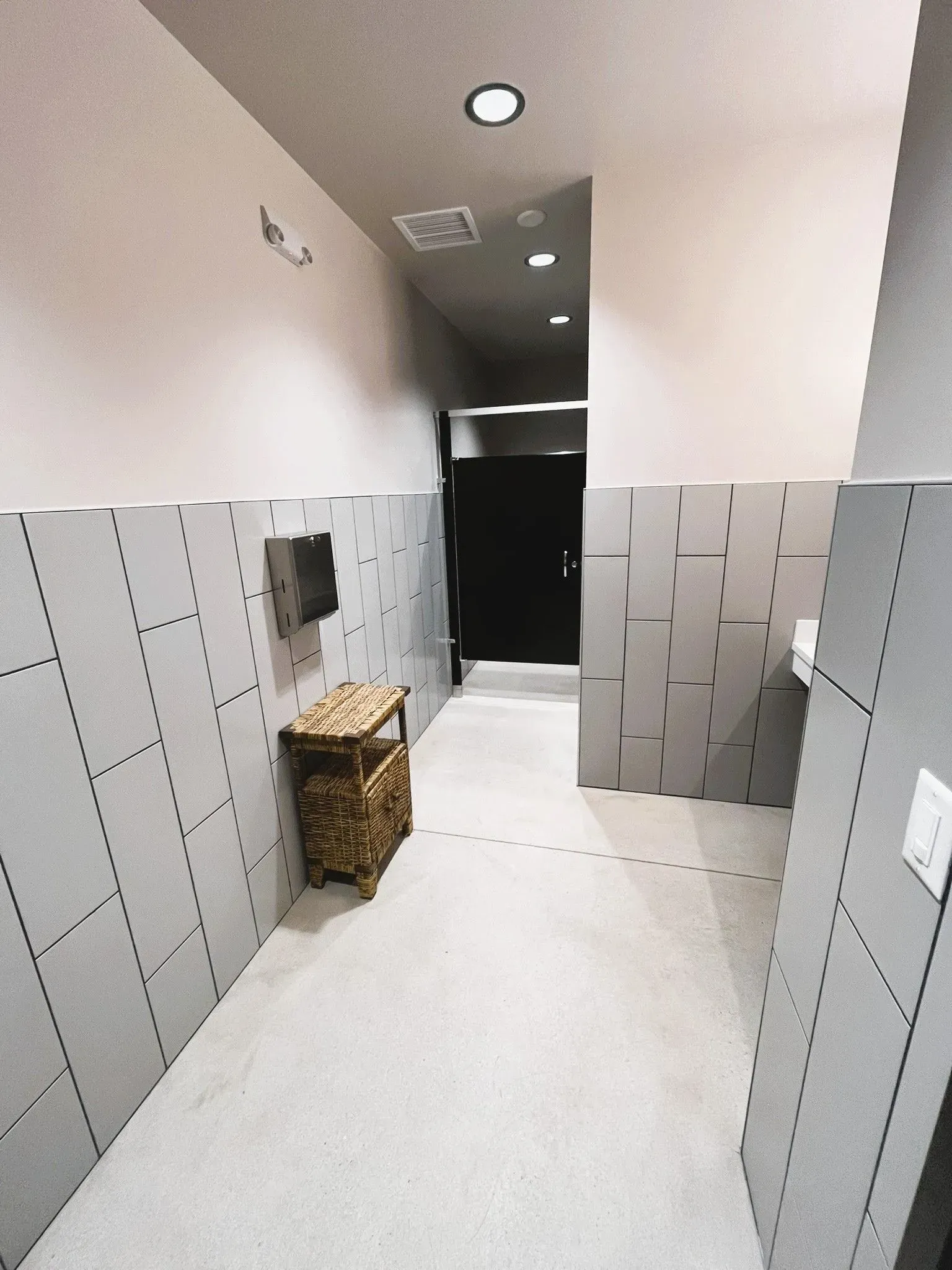 A restroom hallway with light gray walls, dark textured tiling, a small wooden basket, and a dark doorway at the end.