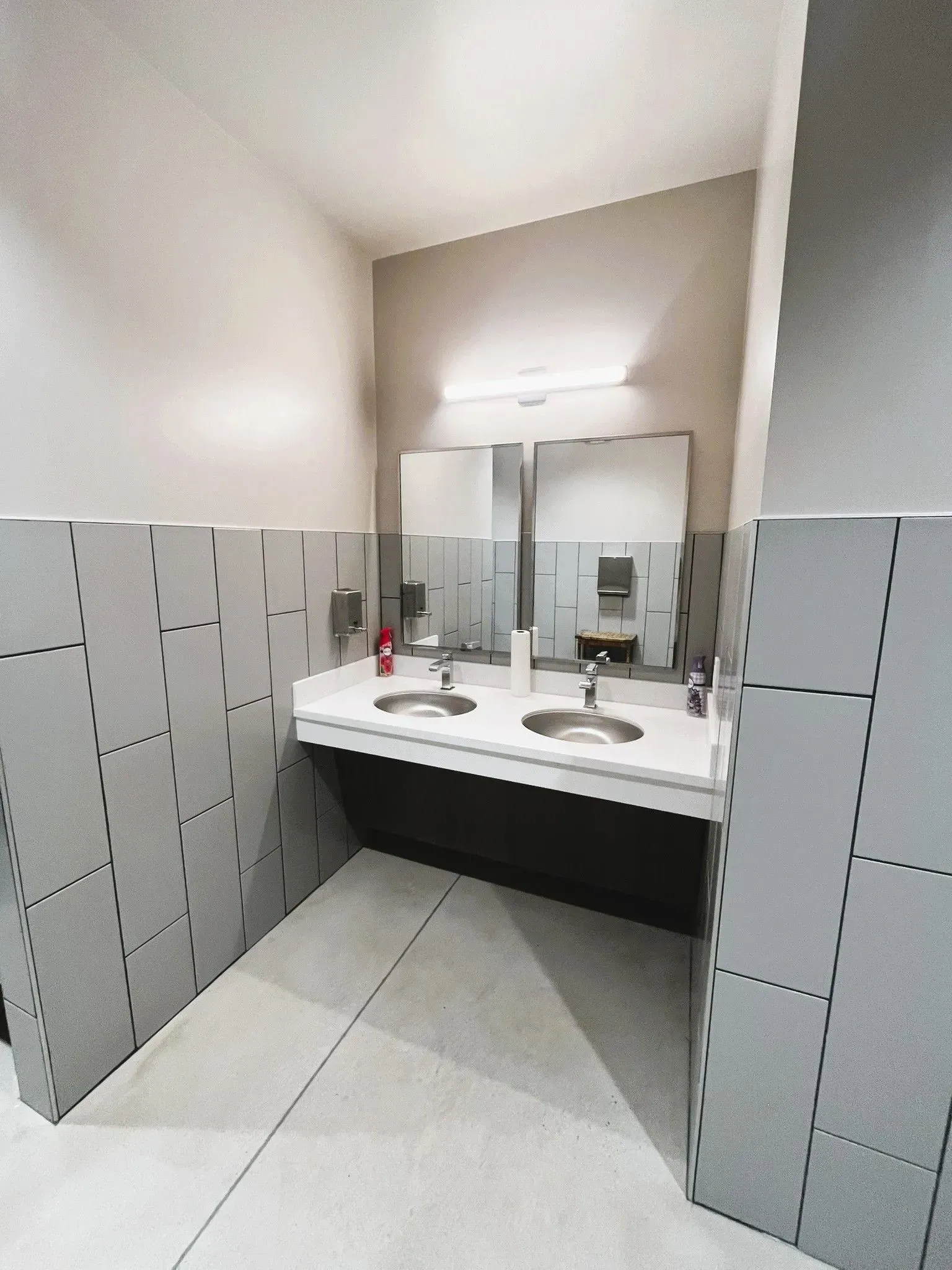 A modern public restroom with two stainless steel sinks set in a white countertop, large mirrors, and gray tiled walls.