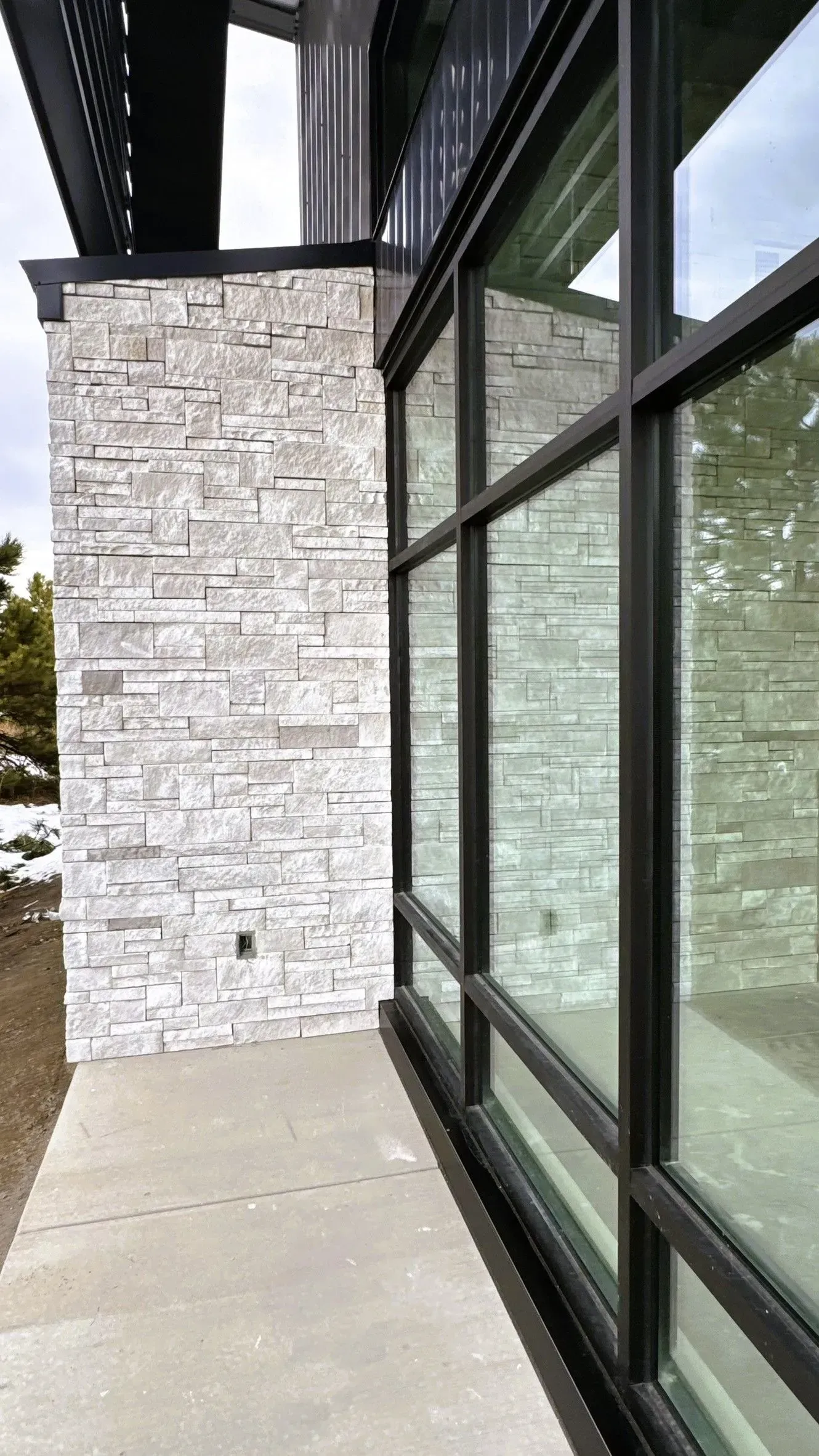 A white stone exterior wall meets a floor-to-ceiling glass window with a black frame on a concrete patio.