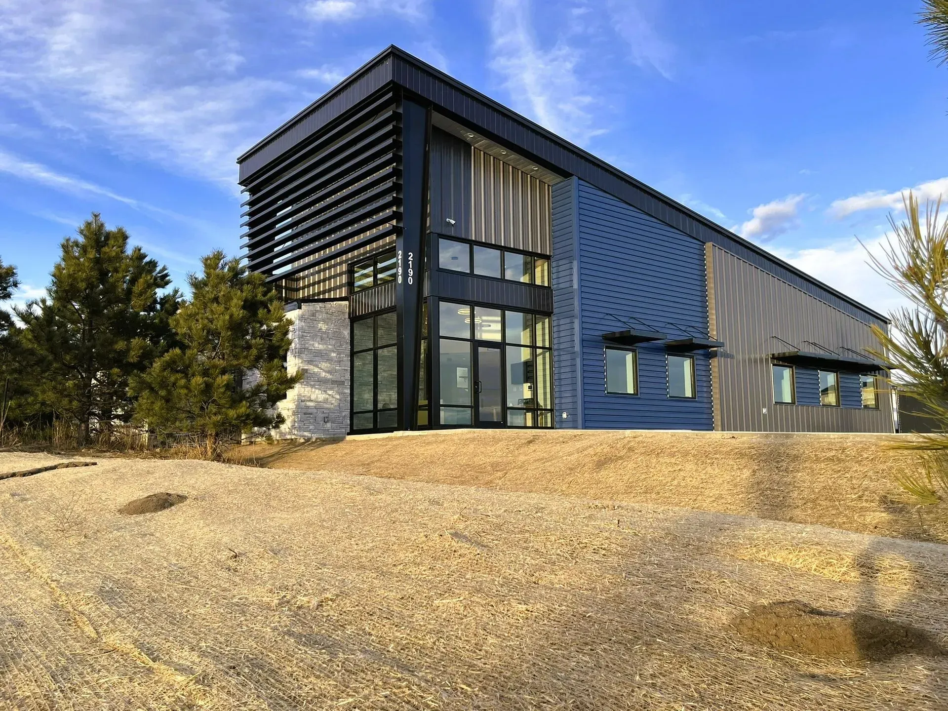 Modern industrial-style building with blue corrugated metal walls, large glass windows, and a sloped roof under a blue sky.