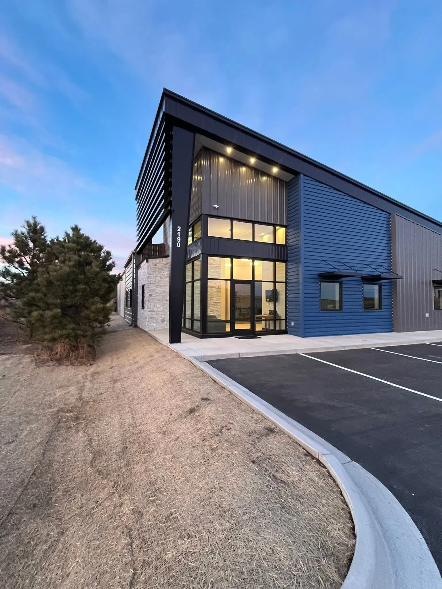 A modern commercial building with blue metal siding, a stone accent wall, and a large glass entryway at dusk.