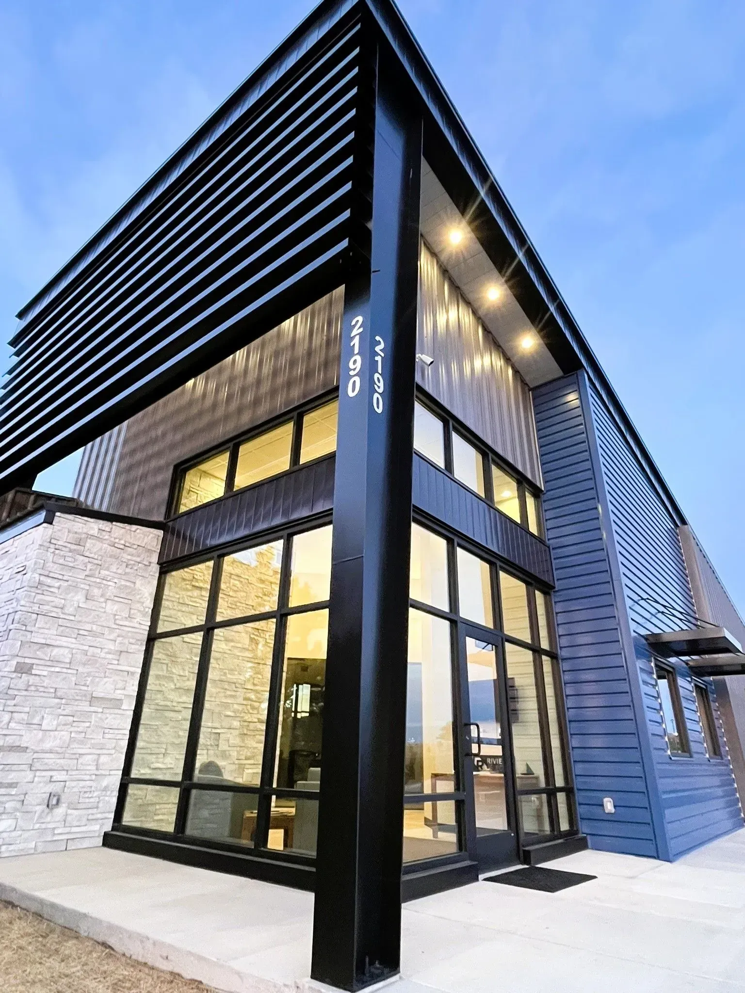 Exterior view of a modern commercial building with a black steel frame, stone walls, and large floor-to-ceiling windows.