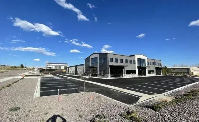 A modern, multi-story grey industrial building under a bright blue sky, featuring a large parking lot in the foreground.