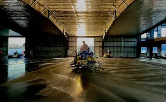A person operates a ride-on concrete power trowel inside a large, open-air industrial building during sunrise.