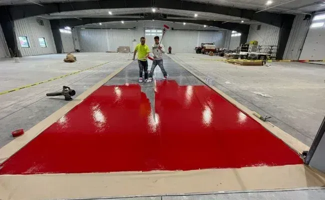 Two workers stand on a concrete floor in a warehouse, overlooking a freshly applied, glossy red epoxy coating strip.