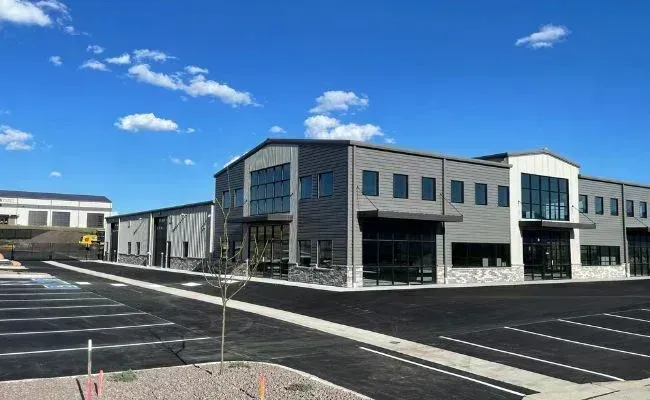 A modern, two-story commercial building with gray siding, glass entryways, and a large, paved parking lot under a blue sky.