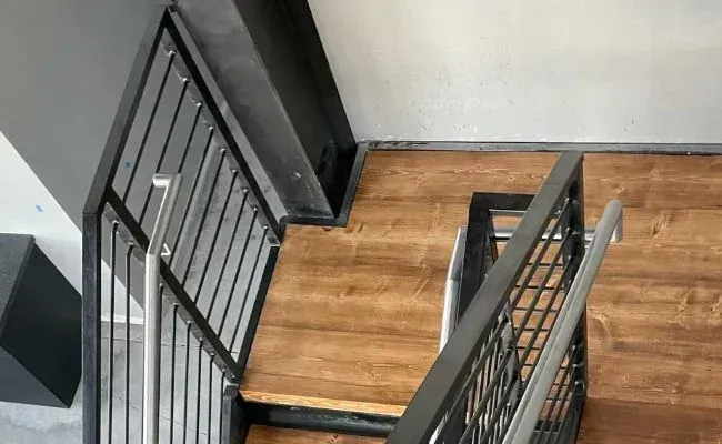 A high-angle view of a wooden staircase featuring a black metal railing and a brushed metal handrail.