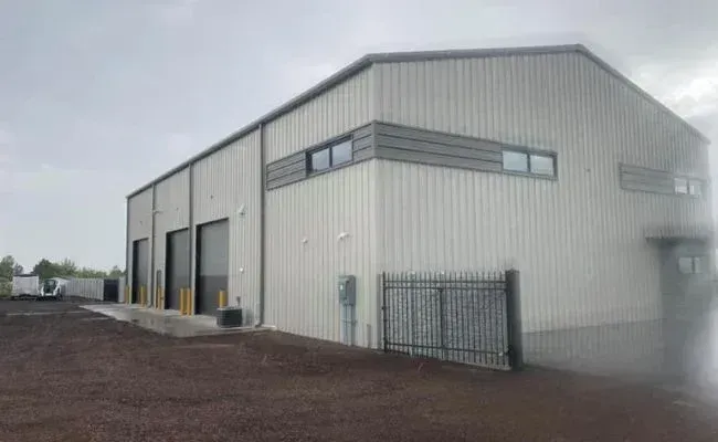 A light-colored metal warehouse building with three garage doors and a fenced side gate on a dirt lot under a cloudy sky.