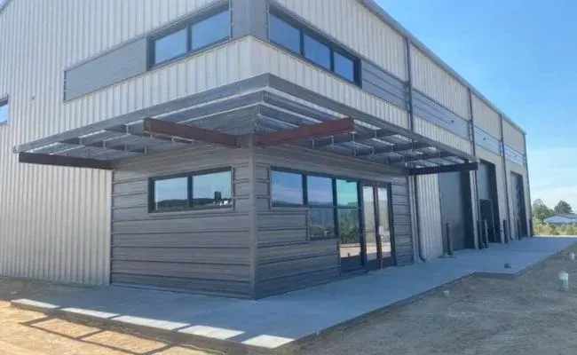 A modern industrial building with horizontal gray siding, a covered entrance, and a concrete patio under a blue sky.