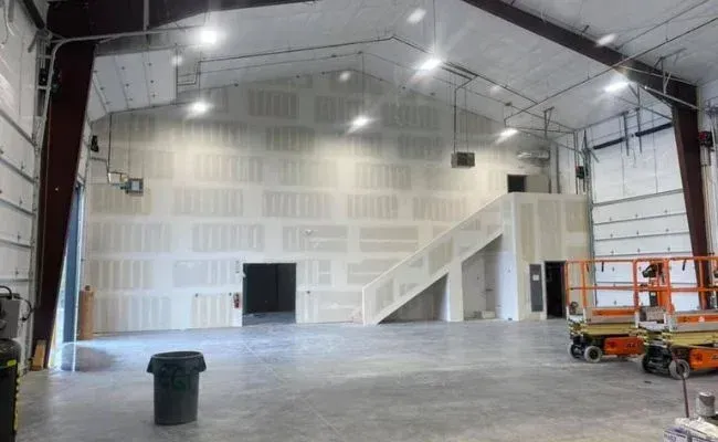 An unfinished warehouse interior featuring drywall panels, a staircase, and construction equipment on a concrete floor.