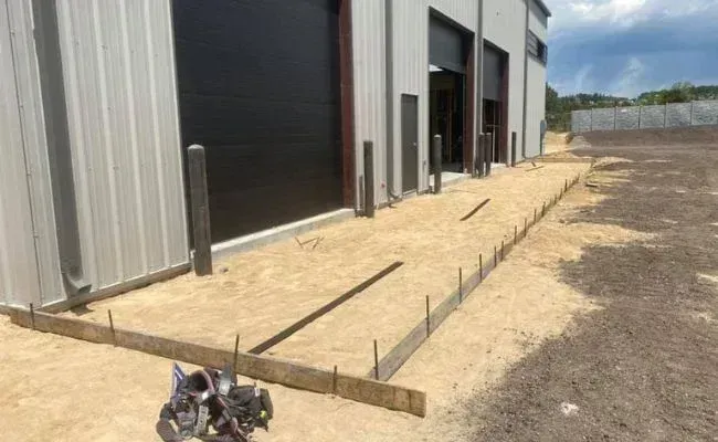 Wooden formwork and steel stakes installed on a sandy ground surface in front of a metal industrial building.