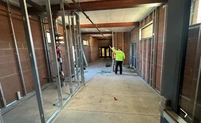 A construction worker in a neon yellow safety jacket walks down an unfinished indoor hallway with metal wall framing.