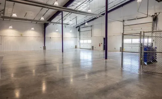 Wide, empty warehouse interior with high ceilings, concrete floors, white walls, and a fenced-in area on the right.
