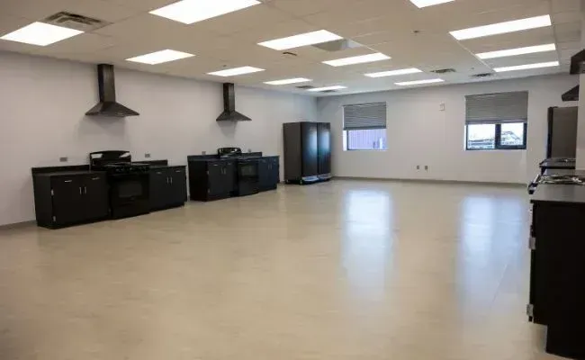 A bright, spacious professional kitchen with black cabinets, stoves, stainless steel hoods, and white walls.