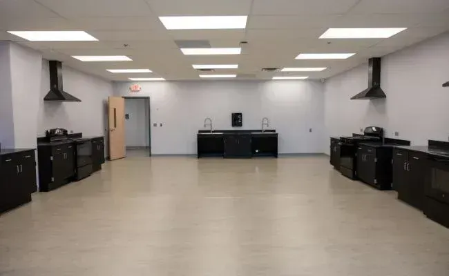 A wide, empty teaching kitchen with light-colored flooring, white walls, black cabinets, stovetops, and exhaust hoods.