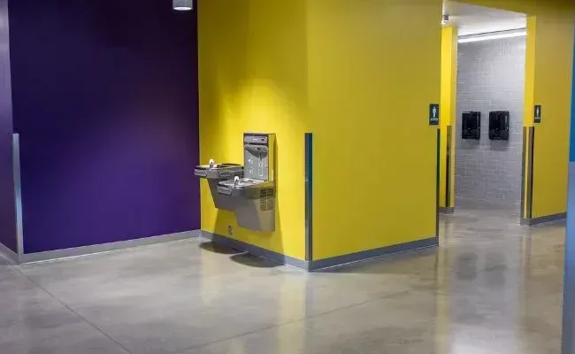 A water fountain mounted on a yellow wall next to a purple wall and a hallway entrance with signage.