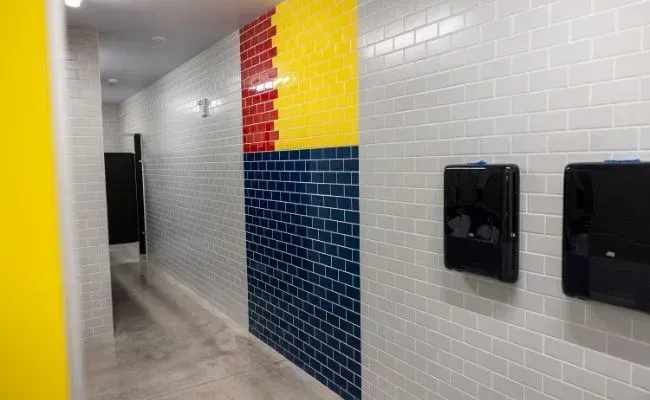A hallway with white subway tile walls, featuring a bold blue, yellow, and red accent section and two black dispensers.
