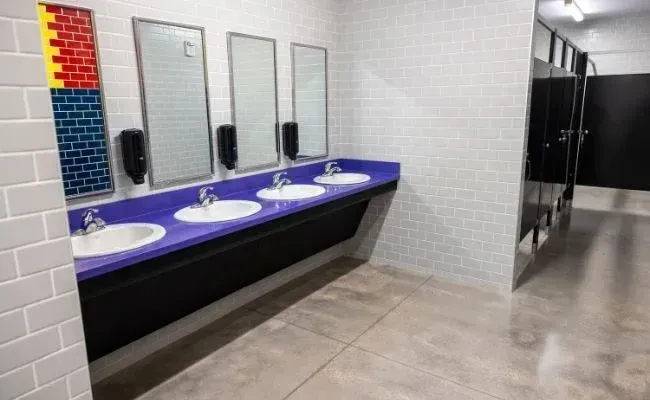 Public restroom with three white sinks on a purple countertop, three mirrors, and black toilet stalls against tiled walls.