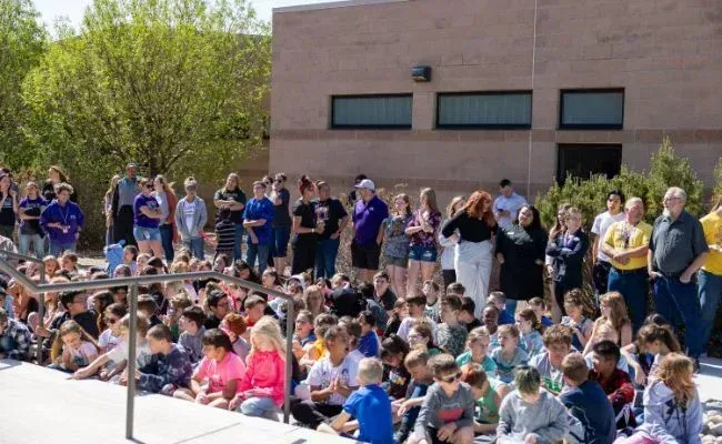 A large group of people gathered outside a brick building, with many sitting on the ground and others standing behind.