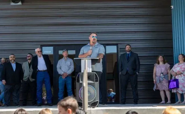 A speaker stands at a podium outdoors in front of a modern building, addressing a group of attendees standing nearby.