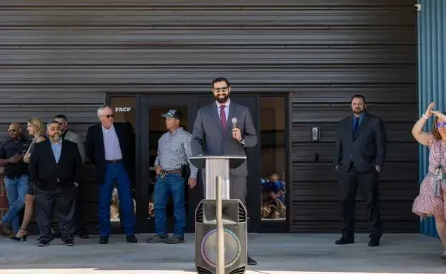 A person in a suit speaks at a podium in front of a modern building, with several people standing nearby.