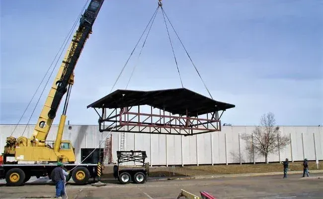 A yellow crane lifts a large, dark metal roof structure in an industrial area under a blue sky.
