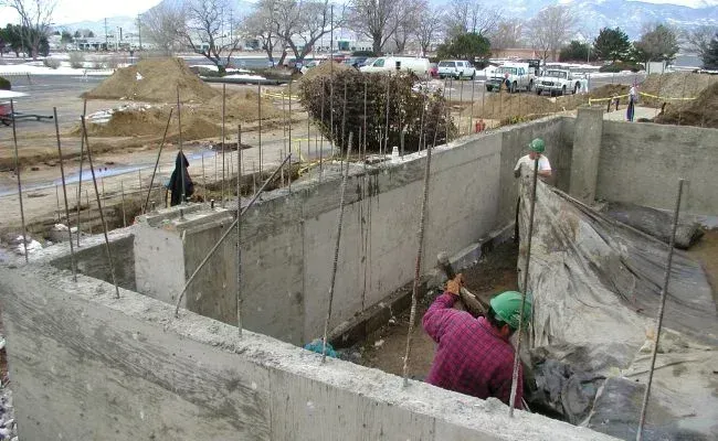 Workers in hard hats build concrete foundation walls on an outdoor construction site with piles of dirt in the background.