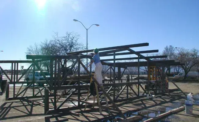 A person stands on a ladder, working on the metal frame of a structure outdoors under a bright, sunny sky.