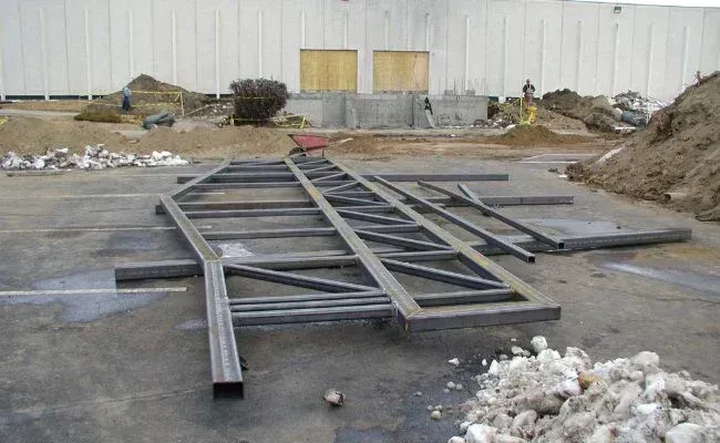 A large, gray steel truss structure lies on a concrete construction site with dirt piles and a building in the background.