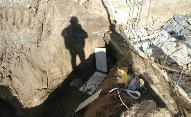 A deep trench excavation site with exposed earth, concrete forms, and equipment, captured under bright daylight.