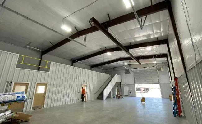 The interior of a high-ceilinged, industrial warehouse with metal walls, a concrete floor, stairs, and a roll-up door.